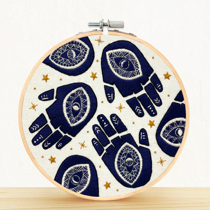 Embroidery hoop with navy blue hand designs and white evil eye details on a beige background with gold stars